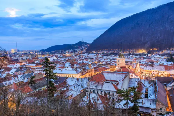 Gökyüzünde dolunay olan bir şehir. Şehir karla kaplı ve binalar aydınlık. 14 Aralık 2024 Brasov Romanya
