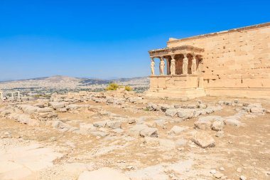Önünde avlu olan büyük bir bina. Avlu boş ve bina kayalarla çevrili. Gökyüzü mavi ve güneş parlıyor, Atina 'da Akropolis