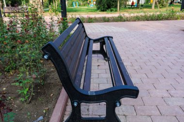 Siyah bir park bankı tuğla bir geçitte oturuyor. Yedek kulübesi boş ve park sessiz, kırık bir element.