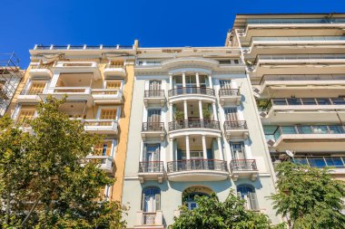 Önünde balkon ve ağaç olan yüksek bir bina. Bina beyaz ve sarı. 8 Ağustos 2024 Selanik Yunanistan