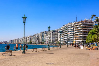 İnsanların banklarda yürüyüp oturduğu bir plaj sahnesi. Sahil yüksek binaları olan bir şehre yakındır. 8 Ağustos 2024 Selanik Yunanistan