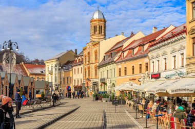 Arka planda büyük bir saat kulesi olan işlek bir şehir caddesi. İnsanlar dışarıda geziyor ve kafelerde oturuyorlar. 14 Aralık 2024 Brasov Romanya