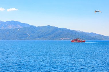 Mavi bir okyanusta kırmızı bir tekne seyrediyor. Gökyüzü açık ve güneş parlıyor. 7 Ağustos 2024 Thassos 'tan Keramoti Yunanistan feribotu