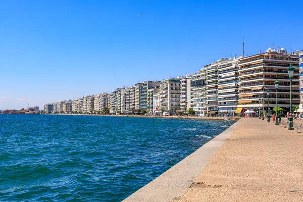 Mavi okyanusu ve binaları olan bir sahil. Binalar yüksek ve balkonları var. Sahil insanlarla dolu ve oturabilecekleri banklar var. 8 Ağustos 2024 Selanik Yunanistan