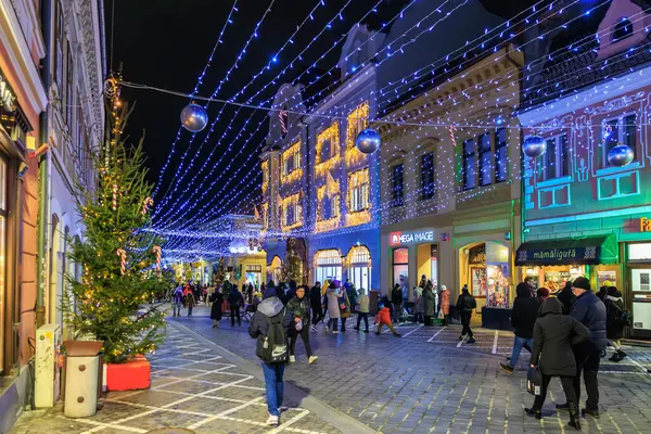 Kalabalık bir cadde, insanlar yürüyor ve önlerinde bir Noel ağacı var. Sokaklar Noel ışıklarıyla aydınlanıyor ve şenlikli bir ortam yaratıyor. 13 Aralık 2024 Brasov Romanya