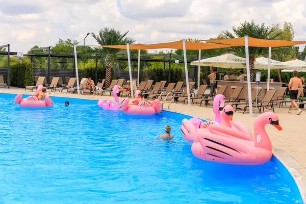 İnsanlar havuzda pembe şişirilebilir yüzdürme cihazları üzerinde yüzüyorlar. Havuz şemsiyeler ve sandalyelerle çevrili ve sandalyelerin üzerinde uzanan birkaç kişi var. 8 Haziran 2024 Balti Moldova