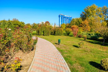 Arka planda mavi bir bina olan bir parktan geçen tuğla bir yürüyüş yolu. Park ağaçlar ve çiçeklerle dolu. Huzurlu ve huzurlu bir atmosfer yaratıyor.