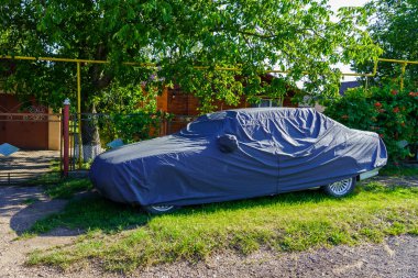 Bir araba siyah bir muşambayla kaplıdır ve çimenli bir alana park edilmiştir. Araba ağaçlar ve çitlerle çevrili.