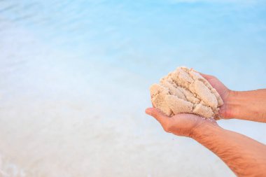 Bir insan kumsalda bir avuç kum tutuyor. Kum beyaz ve su mavi.