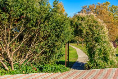 Bir patika büyük bir kemerli bir parktan geçiyor. Kemer yolu ağaçlar ve çalılarla çevrili, huzurlu ve sakin bir atmosfer yaratıyor. Yol tuğla ve taşlardan yapılmıştır.