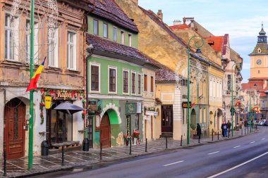 Eski ve yeni binaların karışımından oluşan bir sokak. Binaların çoğu yeşil ve sarı. Sokakta yürüyen birkaç kişi var. 14 Aralık 2024 Brasov Romanya