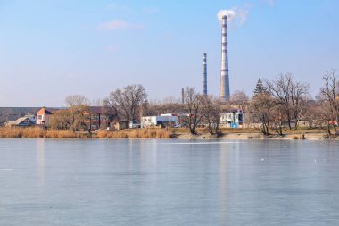 Endüstriyel duman yığınları donmuş göl kenarında çıplak ağaçlarla kaplı kış manzarasında. 22 Şubat 2025 Balti Moldova.