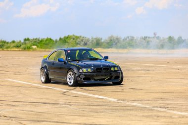 Modifiye edilmiş siyah bir BMW, açık bir pistte dönerken yoğun bir duman izi bırakarak sürükleniyor. 2 Temmuz 2023 Liadoveni Moldova.