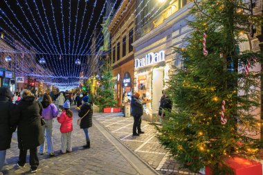 Bir grup insan bir mağazanın önünde elinde Noel ağacıyla caddede yürüyorlar. İnsanlar sıcacık giyinmiş ve festival atmosferinin tadını çıkarıyor gibi görünüyorlar. 13 Aralık 2024 Brasov Romanya