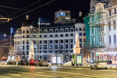 Aydınlatılmış heykelleri ve trafiği olan tarihi şehir mimarisinin gece manzarası. 8 Mart 2025 Bükreş Romanya.