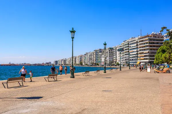 İnsanların banklarda yürüyüp oturduğu bir plaj sahnesi. Sahil yüksek binaları olan bir şehre yakındır. 8 Ağustos 2024 Selanik Yunanistan