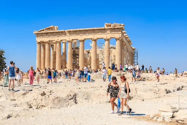 Büyük bir grup insan büyük bir binanın etrafında yürüyor. İnsanlar kuyrukta yürüyorlar ve bazıları sırt çantası taşıyor. 9 Ağustos 2024 Atina Yunanistan