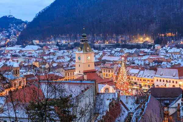 Geceleri ortasında uzun bir saat kulesi olan bir şehir. Şehir karla kaplı ve binaların ışıkları sıcak ve rahat bir atmosfer yaratıyor. 14 Aralık 2024 Brasov Romanya