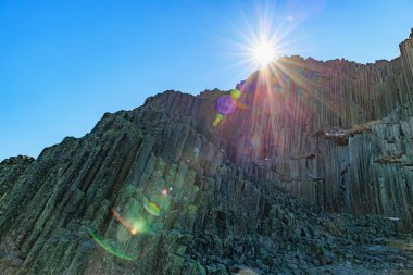 Parlak mercekli güneş ışığı bazalt sütunları açık mavi gökyüzüne karşı parlıyor.