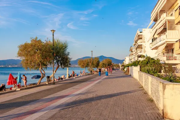 İnsanların tuğla kaldırım boyunca yürüdüğü bir plaj sahnesi. Gökyüzü mavi ve su sakin. 8 Ağustos 2024 Perea Yunanistan