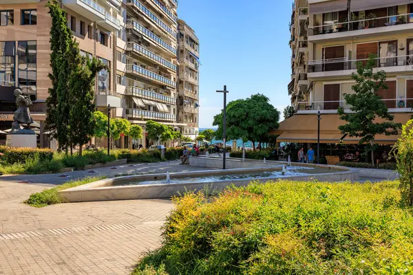 Ortasında çeşme olan bir şehir caddesi ve solunda bir adamın heykeli. Sokak binalar ve ağaçlarla dolu. 8 Ağustos 2024 Selanik Yunanistan