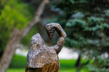 Ahşap heykel, yemyeşil parkta üzüm tutan pelerinli bir figür. 2 Mayıs 2023 Romanesti Moldova.