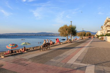 İnsanların banklarda ve şemsiyelerin altında oturduğu bir plaj sahnesi. Olay yeri sakin ve sakin bir yer. 8 Ağustos 2024 Perea Yunanistan