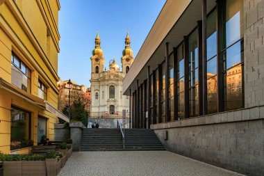 Avrupa 'nın bir şehrinde modern şehir mimarisi tarafından çerçevelenmiş tarihi kilise cephesi. 18 Mart 2025, Karlovy Vary, Çek Cumhuriyeti.