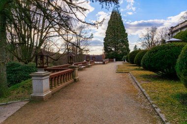 Mavi gökyüzünün altındaki bankların ve ağaçların bulunduğu sakin parktaki manzara yolu. 17 Mart 2025, Konopiste Şatosu, Çek Cumhuriyeti.