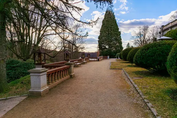 Mavi gökyüzünün altındaki bankların ve ağaçların bulunduğu sakin parktaki manzara yolu. 17 Mart 2025, Konopiste Şatosu, Çek Cumhuriyeti.