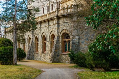 Ağaçlar ve kemerli pencerelerle çevrili tarihi taş bina.