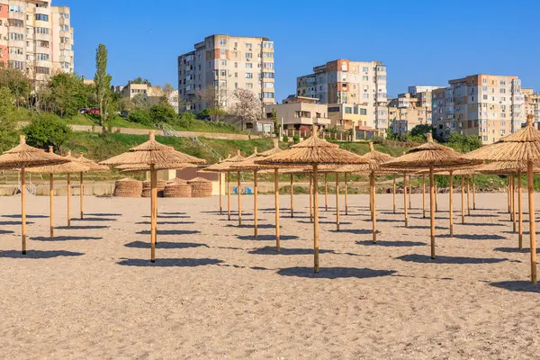 Arkasında saman şemsiyeler ve şehir manzarası olan güneşli bir sahil. 4 Mayıs 2025 Romanya.