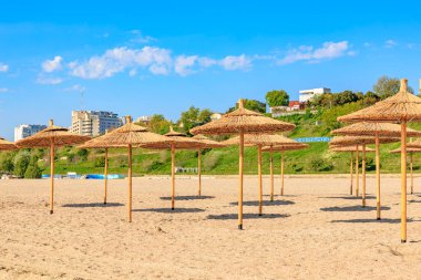 Arkasında saman şemsiyeler ve binalar olan güneşli bir sahil. 4 Mayıs 2025 Romanya.