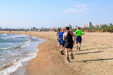 Arka planda şehir manzarası olan bir grup insan plajda koşuyor. 4 Mayıs 2025 Romanya.
