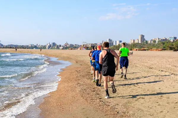 Arka planda şehir manzarası olan bir grup insan plajda koşuyor. 4 Mayıs 2025 Romanya.