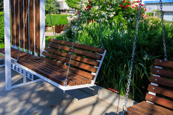 Güneşli bir günde, yemyeşil ve çiçeklerle dolu bir bahçede ahşap veranda salıncak. 5 Haziran 2025 Balti Moldova.