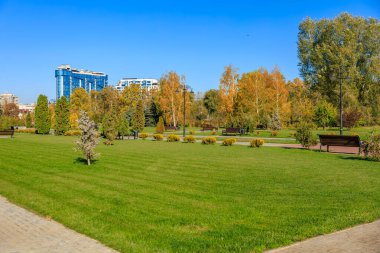 Bir sürü ağaç ve bankların olduğu bir park. Park binalarla çevrilidir. Gökyüzü mavi ve berrak