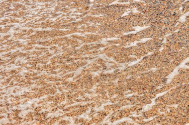 Kumlu sahilde yumuşak dalgalar ve çakıl taşlarında köpüklü su desenleri.