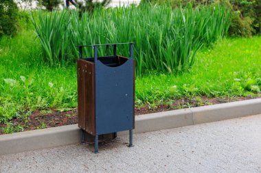Şehir kaldırımındaki yemyeşil çalıların yanında metal çöp tenekesi var..