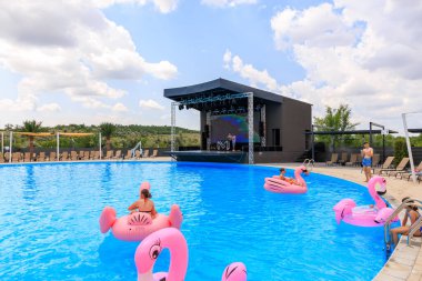 Bir grup insan havuzda şişme flamingo şeklinde yüzüyorlar. Havuz sandalyeler ve şemsiyelerle çevrili ve arka planda bir sahne var. 8 Haziran 2024 Balti Moldova
