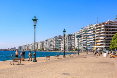 İnsanların banklarda yürüyüp oturduğu bir plaj sahnesi. Sahil yüksek binaları olan bir şehre yakındır. 8 Ağustos 2024 Selanik Yunanistan