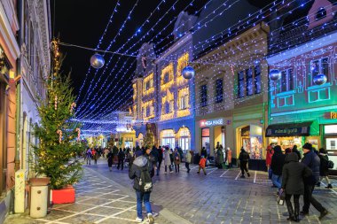Kalabalık bir cadde, insanlar yürüyor ve önlerinde bir Noel ağacı var. Sokaklar Noel ışıklarıyla aydınlanıyor ve şenlikli bir ortam yaratıyor. 13 Aralık 2024 Brasov Romanya