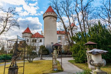 Güneşli bir günde etrafı heykeller ve ağaçlarla çevrili kırmızı taretleri olan tarihi bir şato. 17 Mart 2025, Konopiste Şatosu, Çek Cumhuriyeti.