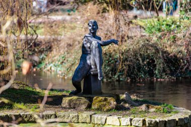 Göletin yakınındaki yemyeşil bahçede oturan bronz heykel. 19 Mart 2025, Karlovy Vary, Çek Cumhuriyeti.