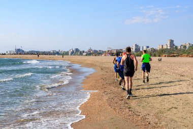 Bir grup genç adam arka planda şehir silüeti olan kumlu sahilde koşuyor. 4 Mayıs 2025 Romanya.