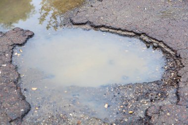 Yıpranmış asfalt yol yüzeyinde sığ su dolu çukurlar..