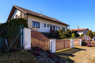 Açık mavi gökyüzünün altında ahşap çitli ve bahçeli bir banliyö evi.