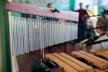 Ahşap tezgahta asılı metal çanlar. Çanlar art arda dizilmiş, bazıları diğerlerinden daha büyük. Sahne bir müzik odasında geçiyor gibi görünüyor.