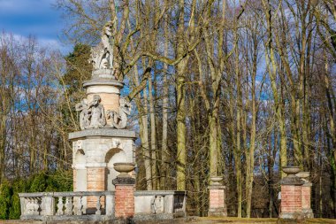Mavi gökyüzünün altında yapraksız ormanda heykelleri olan tarihi taş bir anıt. 17 Mart 2025, Konopiste Şatosu, Çek Cumhuriyeti.