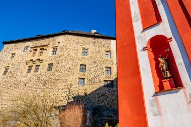 Tarihi taş kale ve güneş ışığında heykeli olan kırmızı duvar. 18 Mart 2025, Madalyon, Çek Cumhuriyeti.